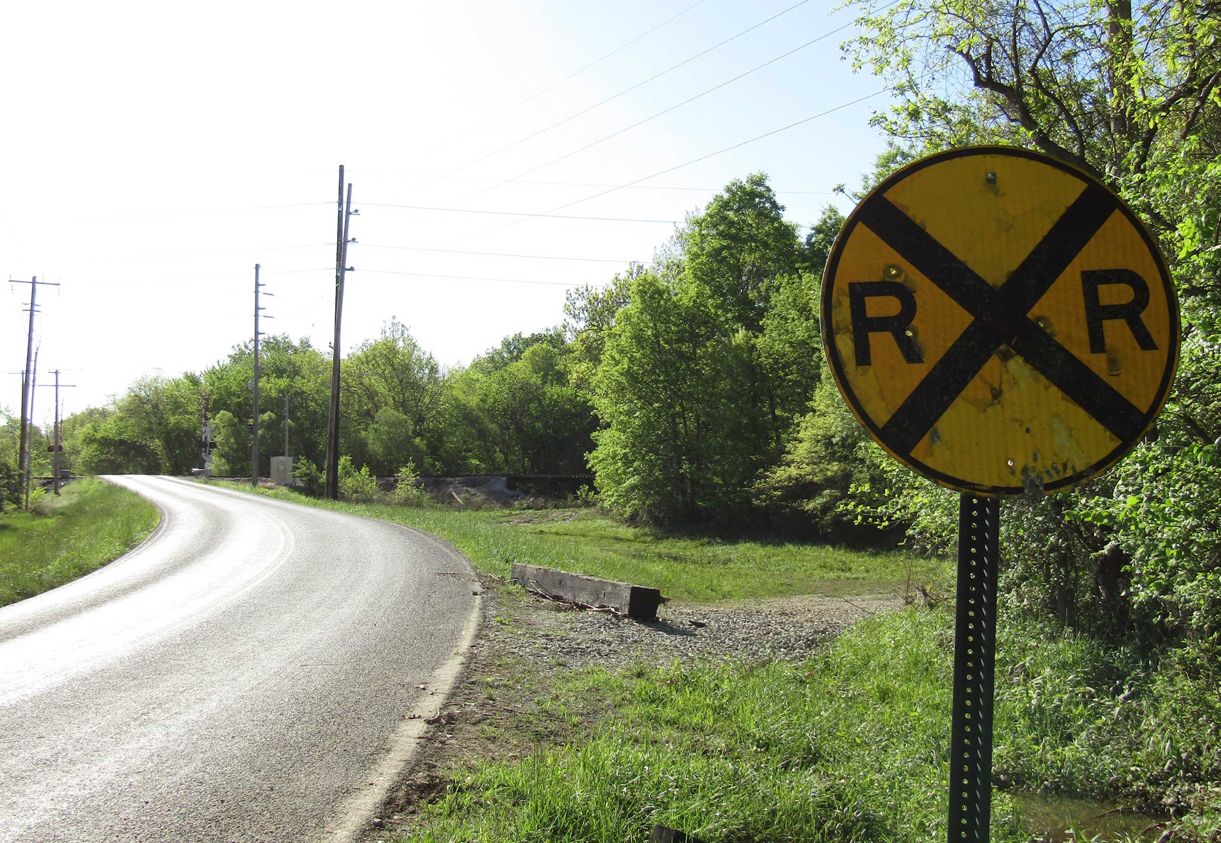 a yellow railroad crossing sign is in the foreground on the right third of the photo, with a gray road curving to the distance on the left side of the photo; in the background are green deciduous trees