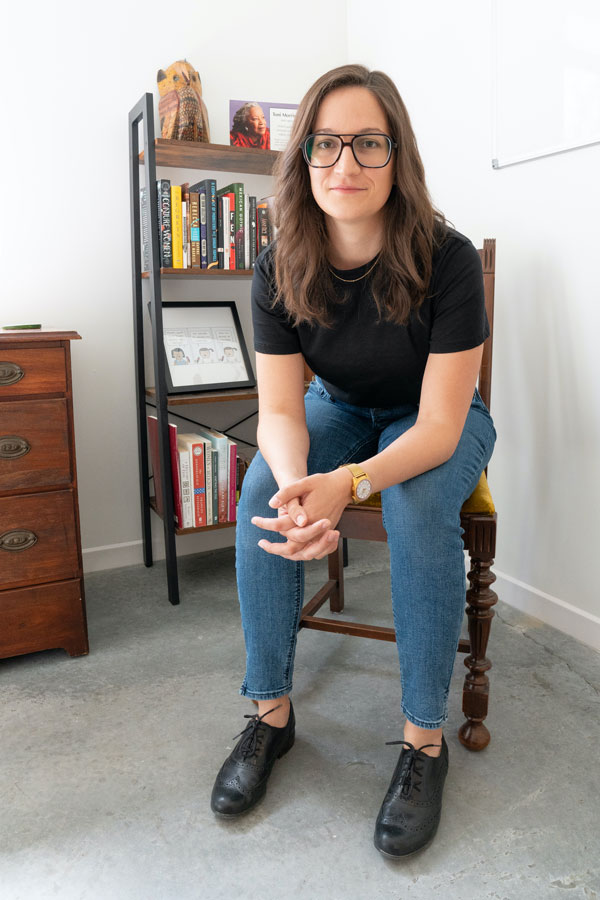 Melanie Pierce sits in a chair in a bright room with a bookshelf behind her left shoulder and a desk on the left side of the portrait. Melanie is white with long brown hair; she wears jeans, a black t-shirt, and black shoes.