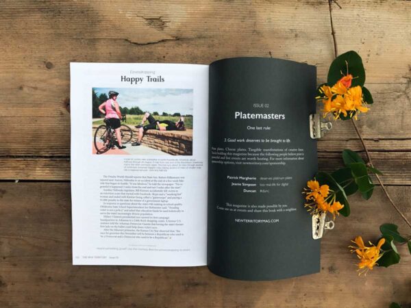 Back page of The New Territory magazine with a photo of cyclists above text on the left-hand side and black inside cover; the magazine is on a warm wood background with yellow honeysuckle placed beside it.
