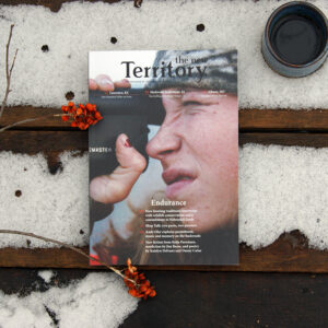 The New Territory issue 08 print magazine rests in the snow with a small mug of coffee to the side and an American bittersweet placed over it. The cover of the magazine depicts a young woman wearing a camouflage cap peering into a handheld hunting scope.