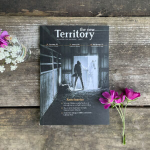 Cover of The New Territory magazine Issue 07: black and white photo by Chase Castor of a man opening the door of a barn to a white horse lays on top of a wood background and pink and white wildflowers