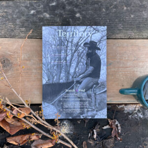 New Territory magazine cover with a cowboy riding a horse. The magazine is sitting on 6" wood slabs and framed by witch hazel and a blue tea mug. The cover photo is black and white.