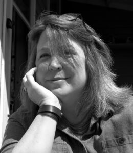 black-and-white portrait of Leslie VonHolten, a white woman with shoulder-length hair and glasses perched on top of her head; she is smiling with her palm under her chin