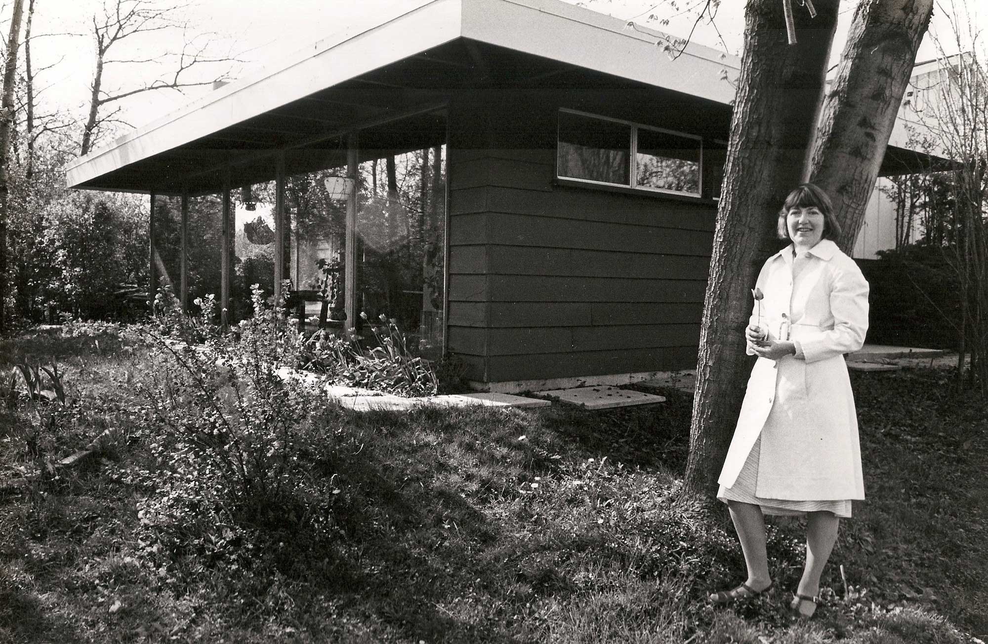Lisel Mueller wearing a white dress outside a new addition to a house. Black-and-white photo.