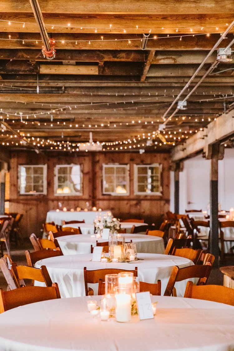 interior hayloft venue with string lights across wood rafters and simple candlelight setting on white tablecloths with wood chairs