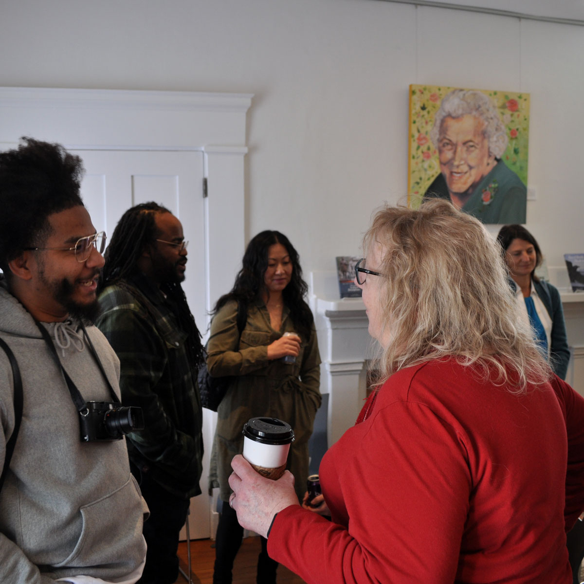 people gathered at an event in a white room with a portrait on the wall. In the foreground two people are conversing: a Black man with an afro and a camera around his neck and a white woman with shoulder-length blond hair, and holding a coffee cup.