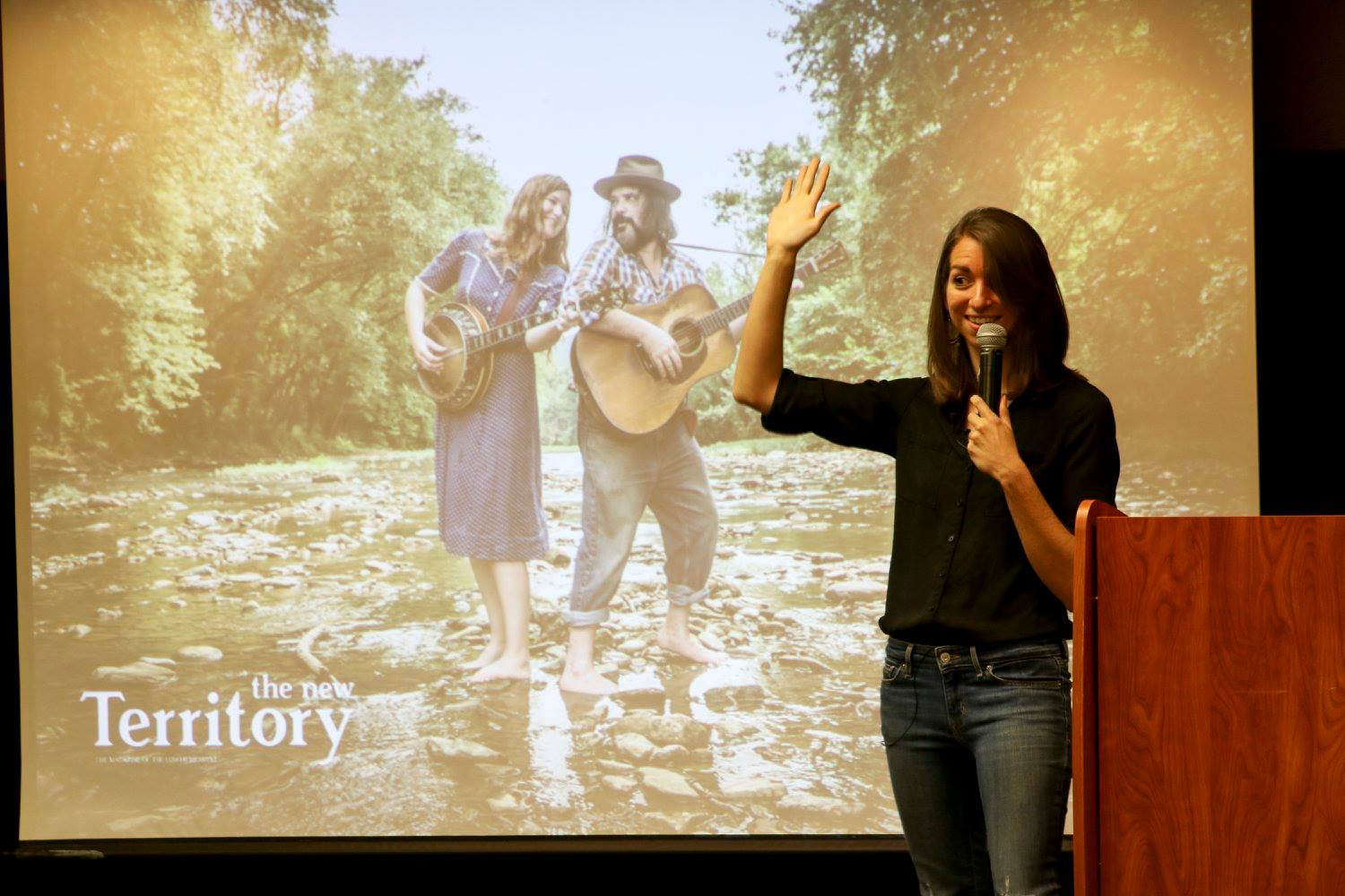 woman holding hand up in front of a screen and podium. The woman is white with brown shoulder-length hair and is wearing jeans and a black button-up shirt. The screen has a two-person string band playing in a creek.