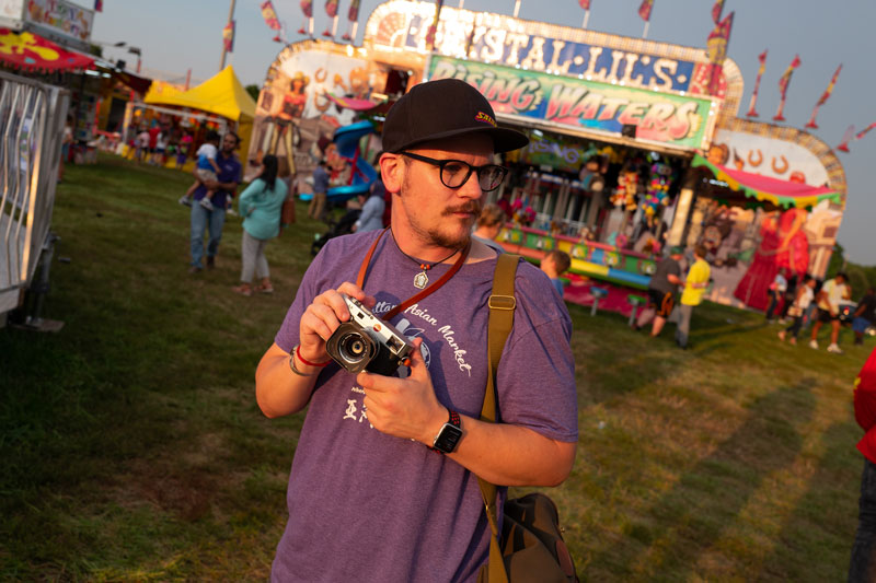 Photographer at a fair. The photographer is white with a purple t-shirt, black hat, and black-rimmed glasses. He holds a camera close to his chest and looks off to his left.