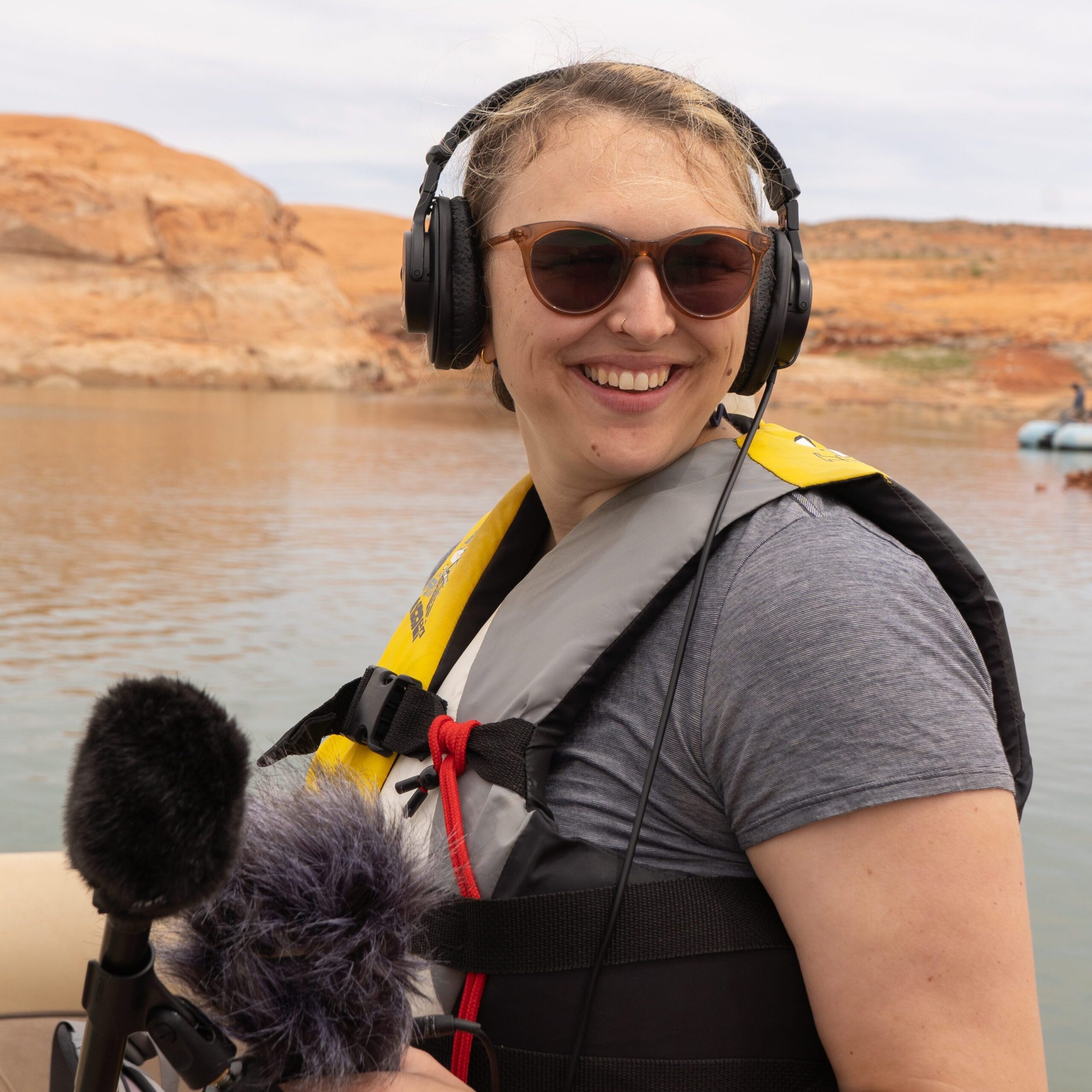 a woman with short, light-colored hair smiles with a microphone and a pair of headphones. The woman is white and the background is a river with red rocks on the banks.