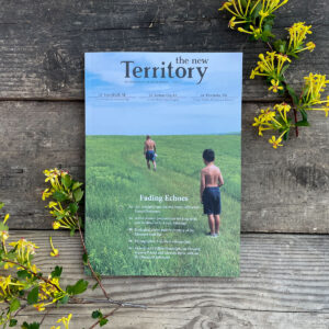 Magazine cover, which features The New Territory nameplate, "Fading Echoes" title, and a photo of a man and a boy in a vast prairie. Magazine is on wood panels and is framed by yellow golden currant flowers.