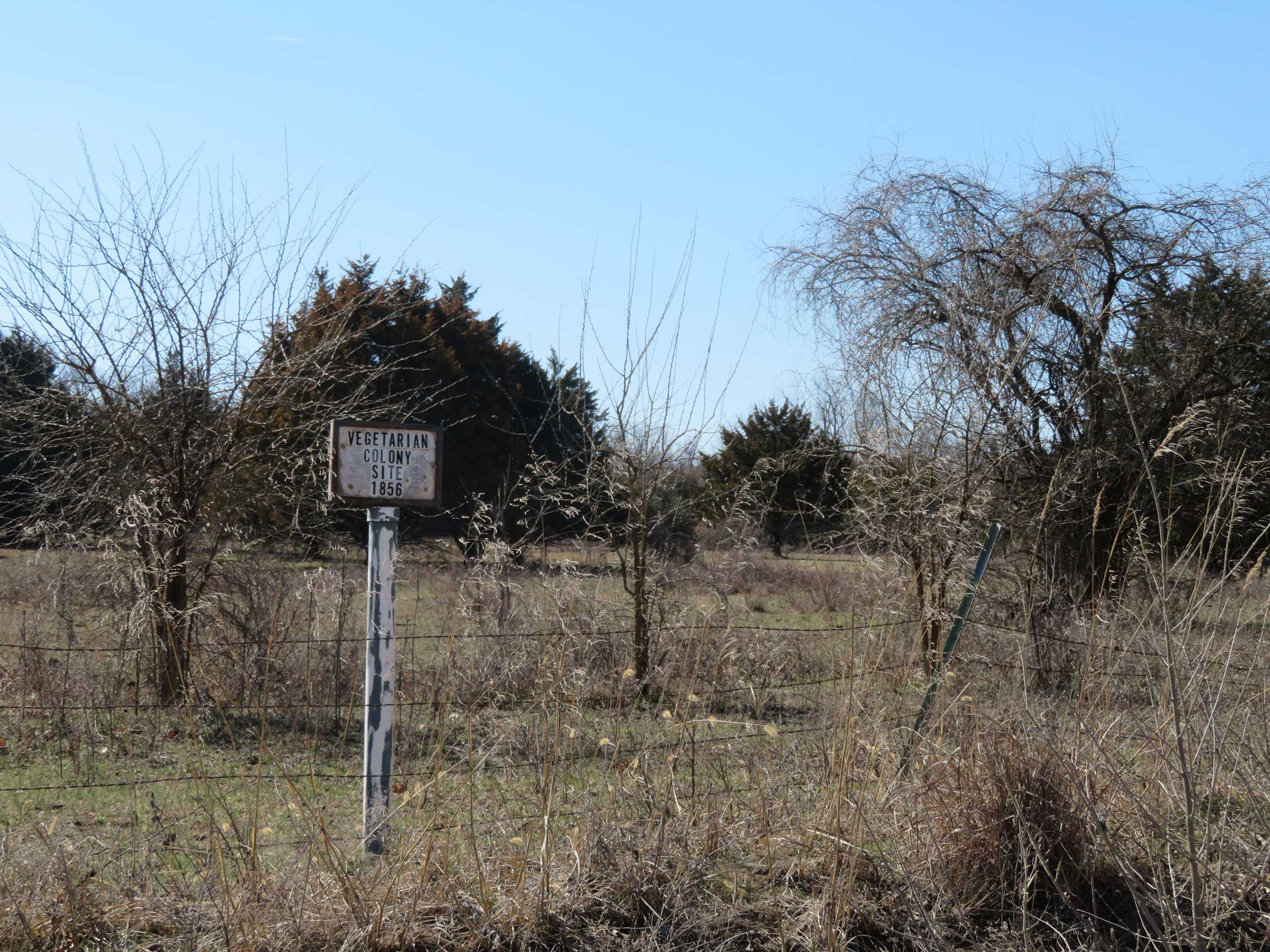 fencerow with small, homemade-looking sign that reads "Vegetarian Colony Site 1856"