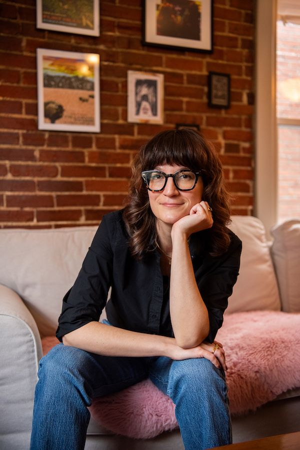 Melanie Pierce sits in a chair in a bright room with a bookshelf behind her left shoulder and a desk on the left side of the portrait. Melanie is white with long brown hair; she wears jeans, a black t-shirt, and black shoes.